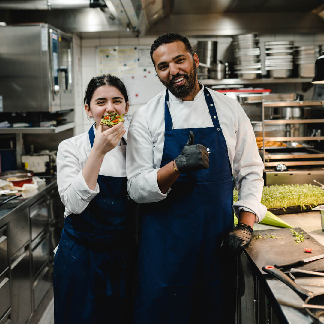 A woman and a man standing in the Restaurant 15 High Kitchen. The man gives a thumbs up and the woman holds some food in her hands.