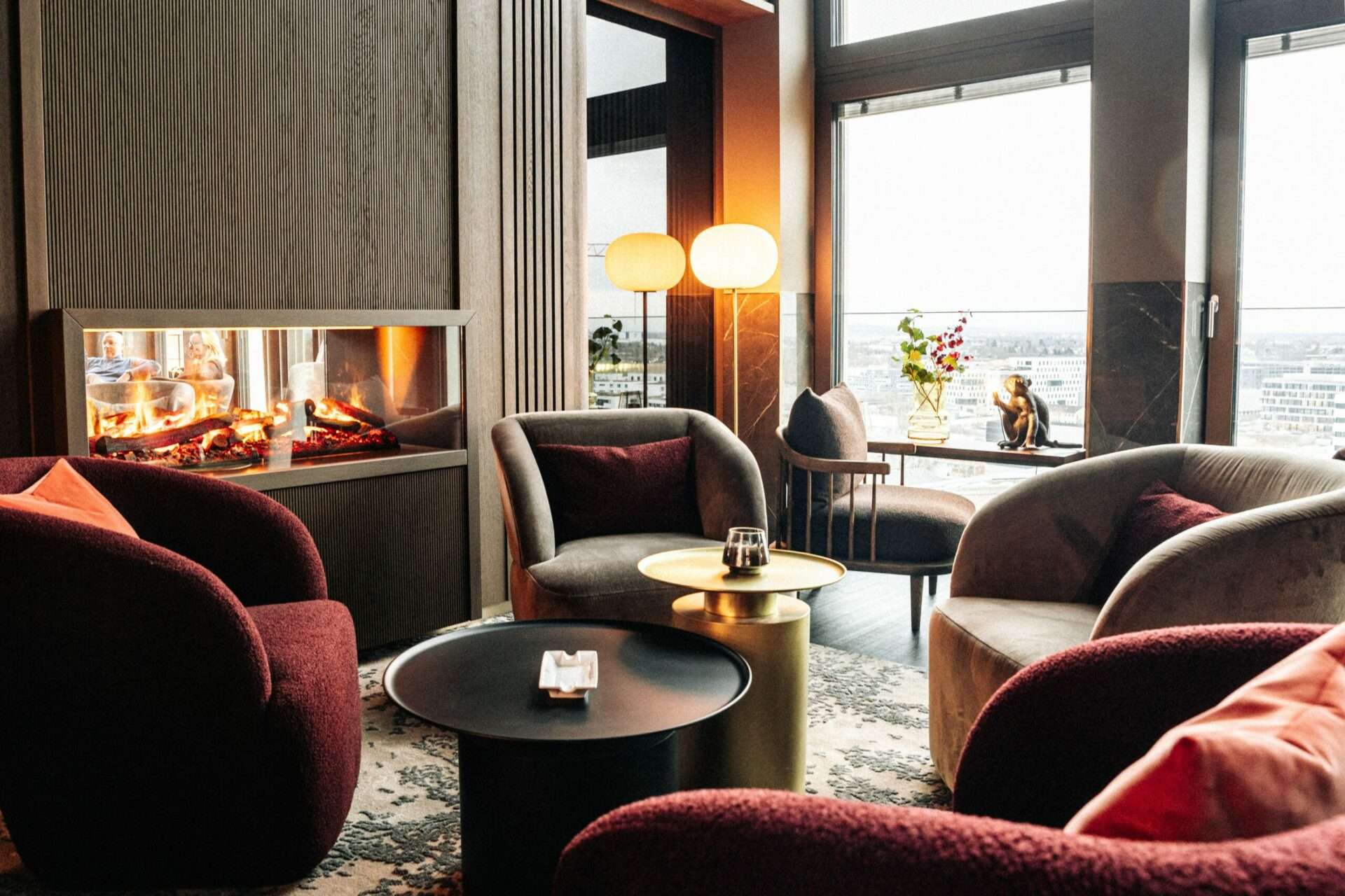 Stools around a small table in front of the fireplace of the 15 High Bar with a view of the city from the window