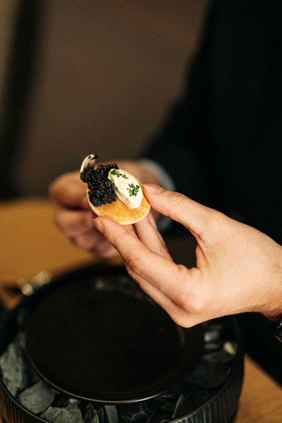 Caviar with buckwheat blini and crème fraîche is held in one hand