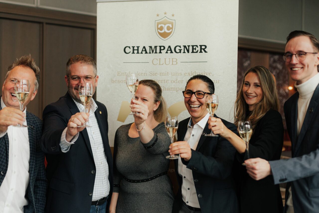 sechs Personen prosten mit einem Champagnerglas in der Hand an